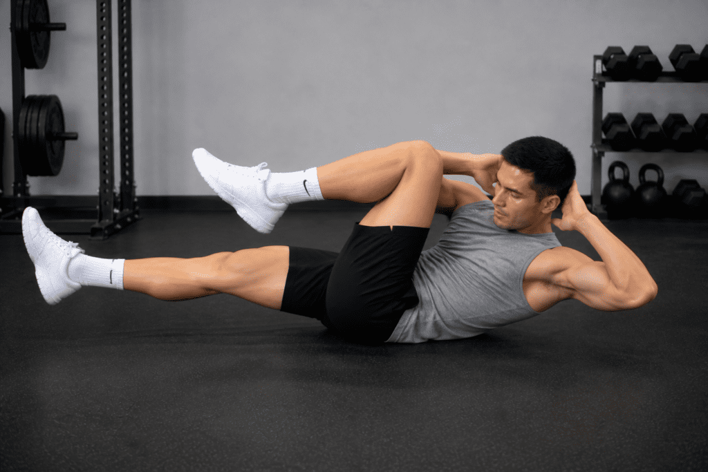 Man in a gray sleeveless shirt performing a bicycle crunch on the gym floor, hands behind head and legs raised.