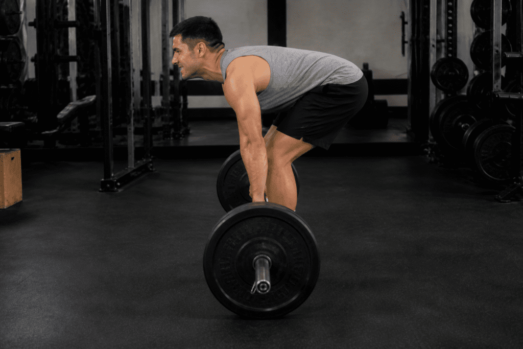 Man in a gray sleeveless shirt and black shorts bent at the hips to lift a loaded barbell in a gym.